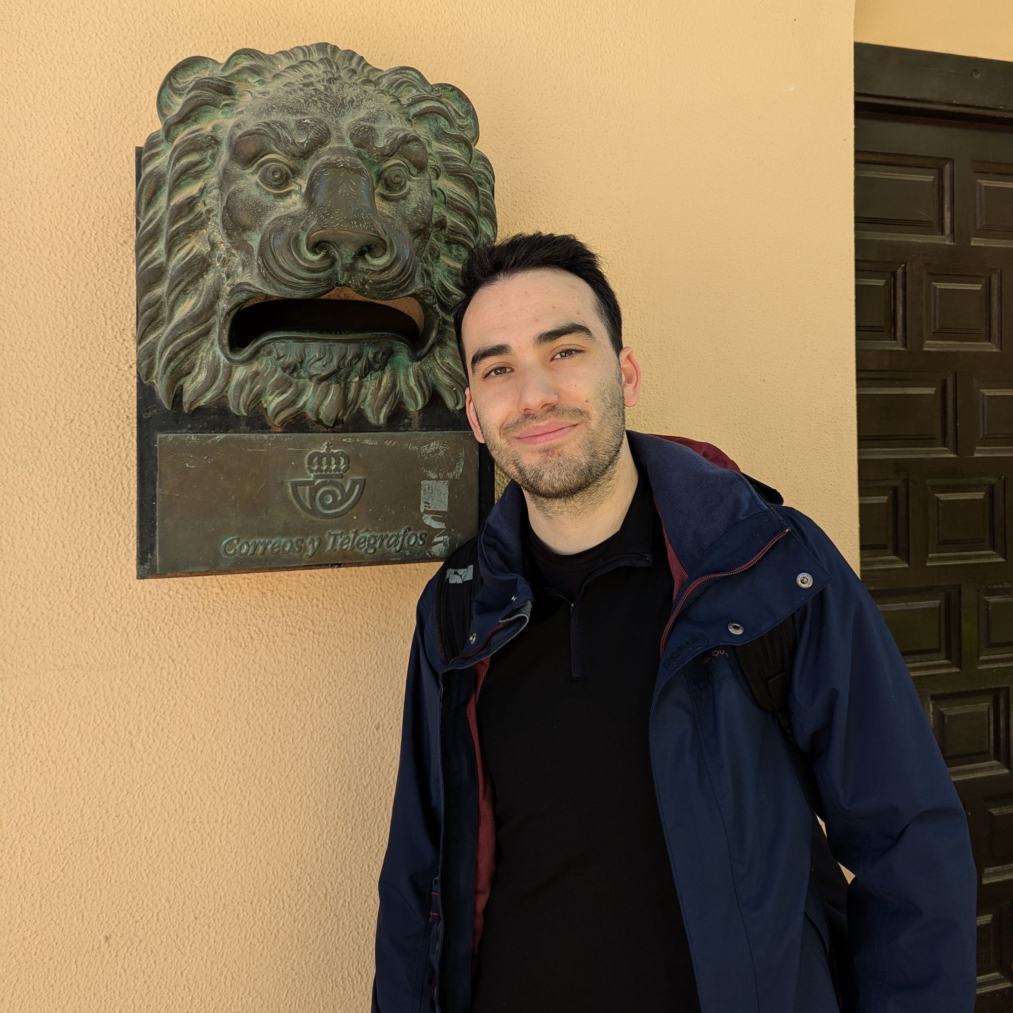 Foto mía, sonriendo delante de un buzón de correos (de los que tienen una cara de León). Llevo un abrigo y una mochila.
